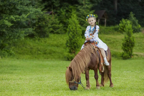 一个身穿乌克兰民族服装的小女孩骑着小马在草坪上 — 图库照片