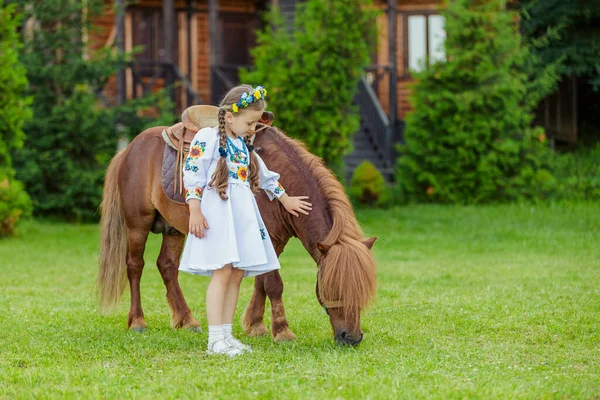 ウクライナの民族衣装の少女は芝生の上で草を食べるポニーをストロークします — ストック写真