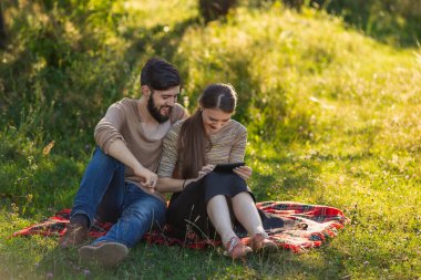 Genç bir çift doğada oturur ve tabletteki bir şeye bakar.