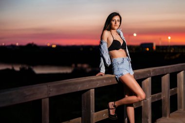 Genç ve seksi bir kadın, şafak vakti denizin yakınındaki tahta bir geçitte poz veriyor..