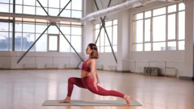 Büyük pencereli bir stüdyoda esneme hareketleri ve yoga egzersizleri yapan atletik bir kız. Asana savaşçısının adı, Virabhadrasana