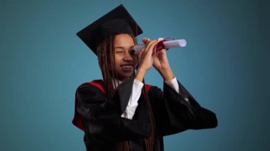 Siyah cüppeli mutlu bir Afrikalı kız ve usta şapkası diplomanın içine bir dürbün gibi bakıyor. Yüksek kalite 4k görüntü