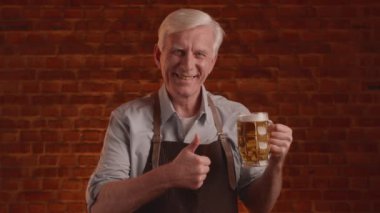 Gri saçlı bira imalatçısı bir bardak taze bira ile poz veriyor. Başparmaklarını gösteriyor, seviyor. Yüksek kalite 4k görüntü