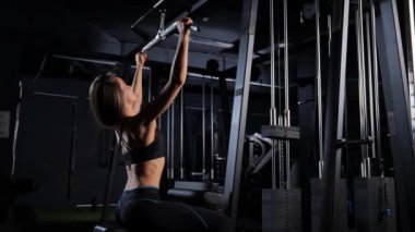 Bir kadın spor salonunda spor aletlerinin üzerine oturmuş, baldır kasları üzerinde çalışıyor, loş ışıklandırma ve flaş fotoğrafa izin verilmiyor.