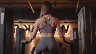 Bir kadın spor salonunda belini, kalçalarını ve karnını yumuşatmak için makineyle çalışıyor. O, mayo ve destekleyici iç çamaşırı giyiyor.