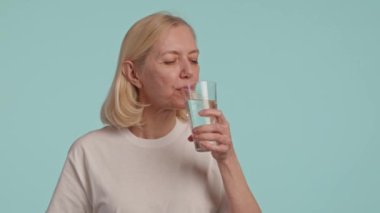 Bir kadın mutlu bir şekilde bir bardak su içiyor, yüzünde bir gülümseme ve kolları havada. Parmağı nazikçe camın kenarına dokunuyor, kirpikleri titreşiyor, sakinleştirici mavi bir arka plana karşı.