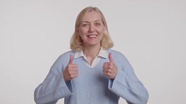 Parlak mavi kazaklı bir kadın gülümsüyor, kolları yanında, mutlu bir jest için iki başparmağını kaldırıyor.