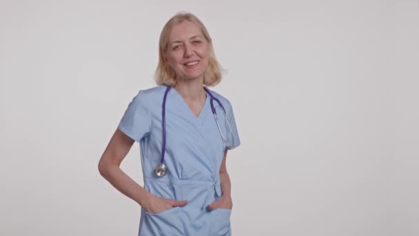 一位穿着蓝色电刷的女医生脖子上戴着听诊器 双手插在口袋里笑着 露出自信的姿势 视频剪辑