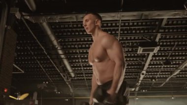 Yalınayak bir adam spor salonunda bir dambılı kaldırarak kaslarını şekillendiriyor ve bir spor şampiyonasına hazırlanıyor.