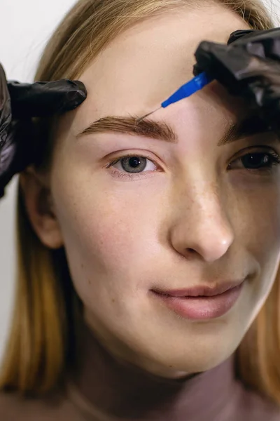 Eyebrow master using a micro brush shapes combing the eyebrows of a red-haired girl with freckles in a beauty salon. Close-up portrait of young woman and hands of stylist in gloves