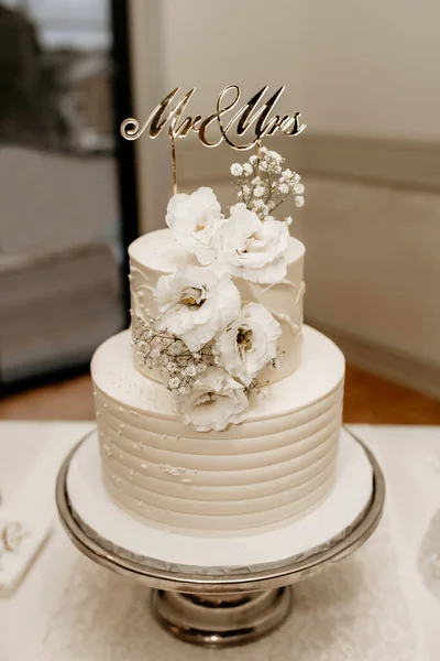 A white wedding cake decorated with flowers and a Mrs. and Mrs. sign are on the table. Beautiful delicious white cake on a silver tray in a restaurant. Wedding cake for newlyweds and their guests