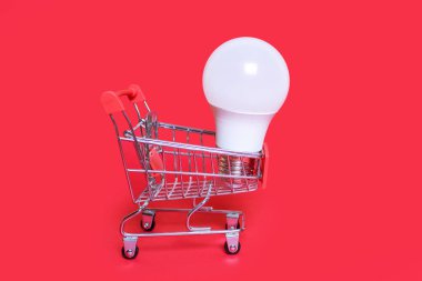 A small shopping cart and an energy-saving light bulb on a red background. Favorable purchase of electric power equipment to save costs. Saving energy and caring for the environment