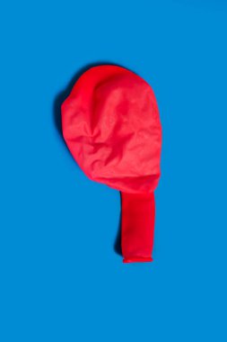 A deflated red balloon on a blue background. One uninflated balloon viewed from above. The concept of the ended holidays in the form of a half-deflated balloon