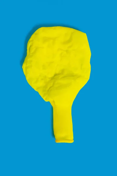 A deflated yellow balloon on a blue background. One uninflated balloon top view. The concept of the ended holidays in the form of a half-deflated yellow balloon
