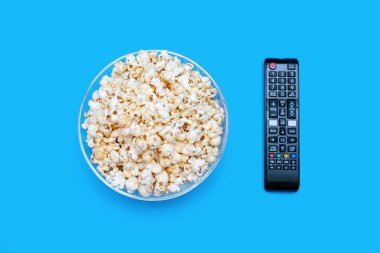 Popcorn in a large glass bowl and a TV remote control on a blue background. A set for watching movies and series in the form of a delicious crispy snack and a TV remote control. Top view of popcorn