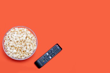 Popcorn in a glass bowl and a TV remote control on an orange background. Leisure time concept with watching movies or series and eating popcorn. Delicious crisp snack. Free space for text