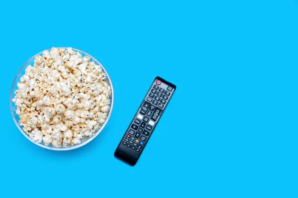 Popcorn in a glass bowl and a TV remote control on a blue background. The concept of watching movies or series with a crispy snack in the form of popcorn. Top view of popcorn. Free space for text
