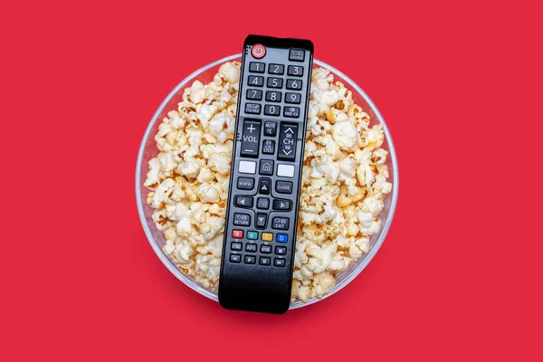 Popcorn in a glass bowl and a TV remote control on a red background. A set for relaxing and watching movies or series. Delicious crispy popcorn snack