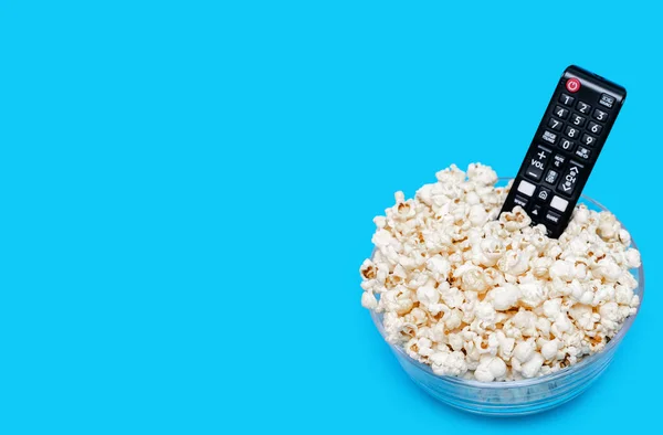 Popcorn in a glass bowl and a TV remote control on a blue background. Leisure time concept with watching movies or series and eating popcorn. View of popcorn and TV remote control. Free space for text