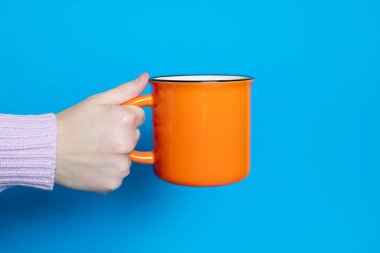 A hand holds a large orange cup with a hot drink on a blue background. The hand of a woman dressed in a pink sweater holds a cup on a blue background. Delicious drink in the form of tea or cocoa