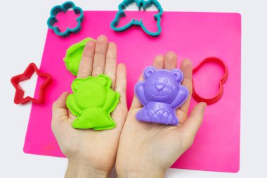 Bir çocuk, yumuşak plastisin ve pembe bir tahta üzerinde modellik yapmak için bir takım kalıplarla oynar. Yakından çekilmiş bir çocuğun ellerinde yeşil havadar plastikler var. Eğlence için çeşitli plastik kalıplar.