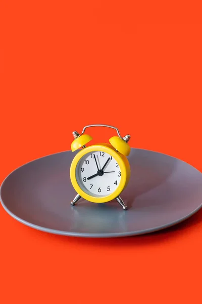 橙色垂直背景上的盘子上竖立着一个黄色的闹钟 饮食和适当营养的概念在时间表上 午餐休息时间或就餐时间 严格按照约定的时间吃饭 — 图库照片