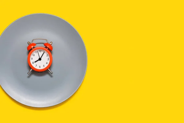 黄色背景上的盘子上有一个红色的闹钟 按时饮食和适当营养的概念 一个明亮的红色闹钟和一个灰色的大盘子 文字和广告的自由空间 — 图库照片