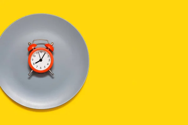 A red alarm clock lies on a plate on a yellow background. The concept of following a diet and proper nutrition on a schedule. A bright red alarm clock and a gray big plate. Free space for text and ads