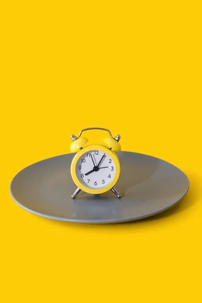 A yellow round alarm clock stands on a plate on a yellow vertical background. The concept of diet and proper nutrition on a schedule. Lunch break or meal time. Eating strictly at the agreed time