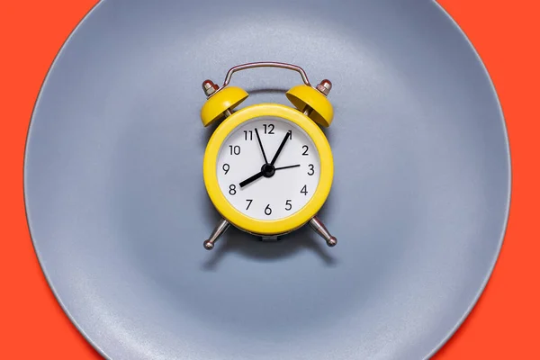 A yellow alarm clock lies on a plate on an orange background. The concept of diet and proper nutrition on a schedule. Lunch break or meal time. Eating strictly at the agreed time