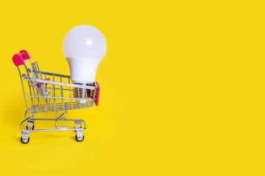 A small shopping cart and an energy-saving light bulb on a yellow background. Favorable purchase of electric power equipment to save costs. Sale of electrical equipment. Free space for text