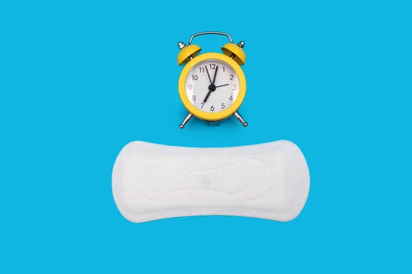 Feminine hygiene menstrual pad for the menstrual cycle and an alarm clock on a blue background. The concept of tracking the menstrual cycle in the form of a watch and a sanitary napkin
