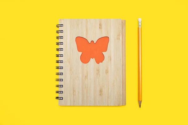 A closed notepad in a bamboo cover with an orange butterfly on a yellow background and a pencil. Stationery in the form of a notepad and a pencil on a yellow background top view