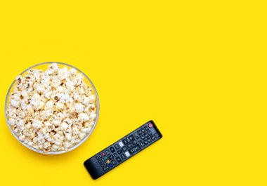 Popcorn in a glass bowl and a TV remote control on a yellow background. Leisure time concept with watching movies or series and eating popcorn. Top view of popcorn. Free space for text