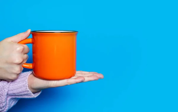 stock image A hand holds a large orange cup with a hot drink on a blue background. The hand of a woman dressed in a pink sweater holds a ceramic cup on a bright blue background. Free space for text