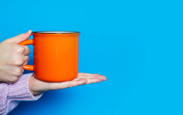 A hand holds a large orange cup with a hot drink on a blue background. The hand of a woman dressed in a pink sweater holds a ceramic cup on a bright blue background. Free space for text