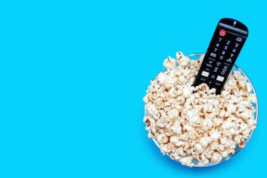Popcorn in a glass bowl and a TV remote control on a blue background. Leisure time concept with watching movies or series and eating popcorn. View of popcorn and TV remote control. Free space for text