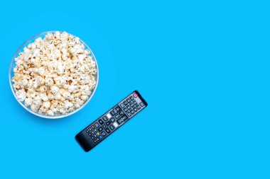 Popcorn in a glass bowl and a TV remote control on a blue background. The concept of watching movies or series with a crispy snack in the form of popcorn. Top view of popcorn. Free space for text