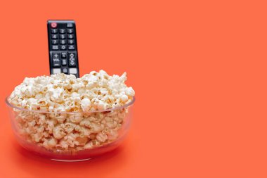 Popcorn in a glass bowl and a TV remote control on an orange background. Leisure time concept with watching movies or series and eating popcorn. Popcorn and TV remote control. Free space for text