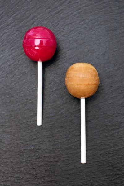 赤とカラメルの2色のロリポップがラフな黒の背景にあります スティック上の古典的な甘いキャンディー 子供のための甘いデザートコンセプト — ストック写真