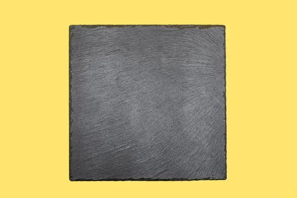 Black slate board in the shape of a square on a yellow background. Empty space for text or design image. Thin slate coaster for cooking or restaurant menus. Free space for text. Black square top view
