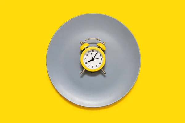 黄色的闹钟停在黄色背景的盘子上 按时饮食和适当营养的概念 亮黄色闹钟和灰色盘子 — 图库照片