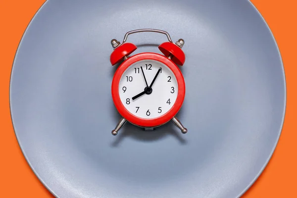 橙色背景的盘子上躺着一个红色的闹钟 饮食和适当营养的概念在时间表上 午餐休息时间或就餐时间 严格按照约定的时间吃饭 — 图库照片