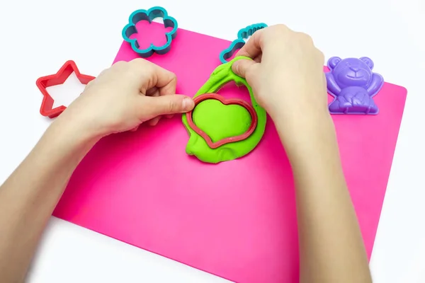 The child plays with soft plasticine and sculpts a green heart using the shape. Hands of a child with airy plasticine and a heart shape close-up. A set of different molds for modeling from plasticine