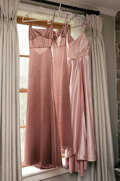 3つの長いピンクの新婦付添人の服は窓にかかっている 美しい新婦付添人の服 スタイリッシュなドレスは部屋に掛けられ 結婚式の準備ができている — ストック写真
