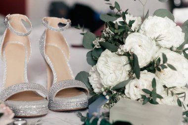 Gelinin yüksek topuklu ayakkabıları ve yakın plan bir gül buketi. Ayakkabı şeklinde düğün aksesuarları ve tören için buket. Gelinin ışıl ışıl ayakkabıları ve lüks bir buket