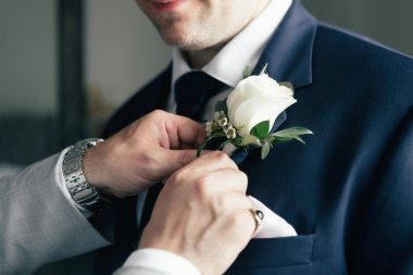 Damadın gül yaka çiçeğini takmasına yardım eden bir arkadaşın yakın çekimi. Damat, düğün töreni için hazırlanıyor ve otel odasında giyiniyor. Damadın arkadaşına yardım et. Gül yaka çiçeği aksesuarı