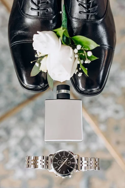 Men's accessories in the form of a mechanical watch, black shoes, perfume and a boutonniere on the wedding day. Groom's accessories top view on the wedding day. White rose boutonniere for the groom