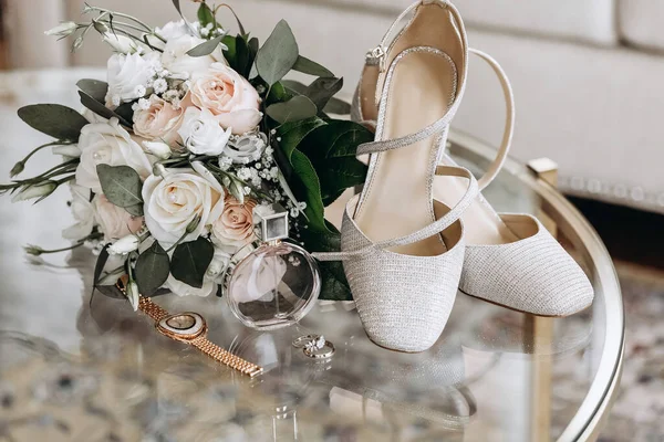 Wedding accessories of the bride in the form of a bouquet of roses, high-heeled shoes, wedding rings and perfume. Bridal bouquet of white roses. Sparkling bride's shoes and gemstone rings close-up