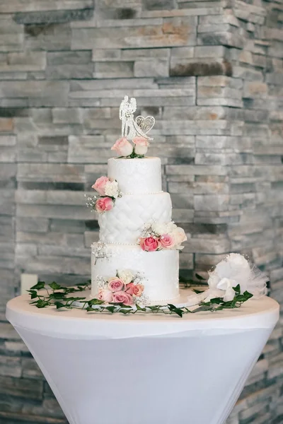 White wedding cake decorated with red roses and figurine in the shape of a bride and groom. Luxurious tiered cake in a restaurant. Traditional wedding cake on a vertical image